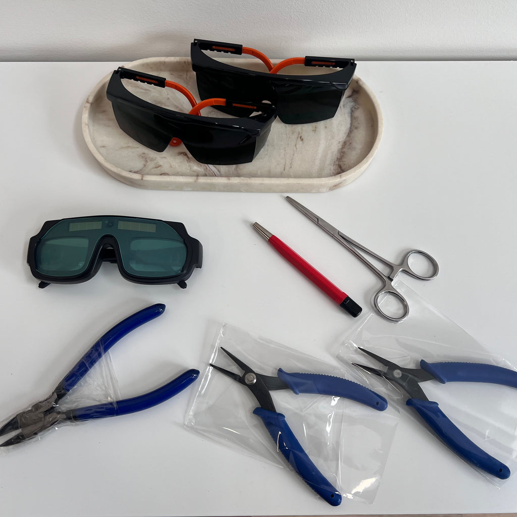 Pair of safety goggles, pliers, scissors, and a pen on a white surface with a marble-patterned tray.