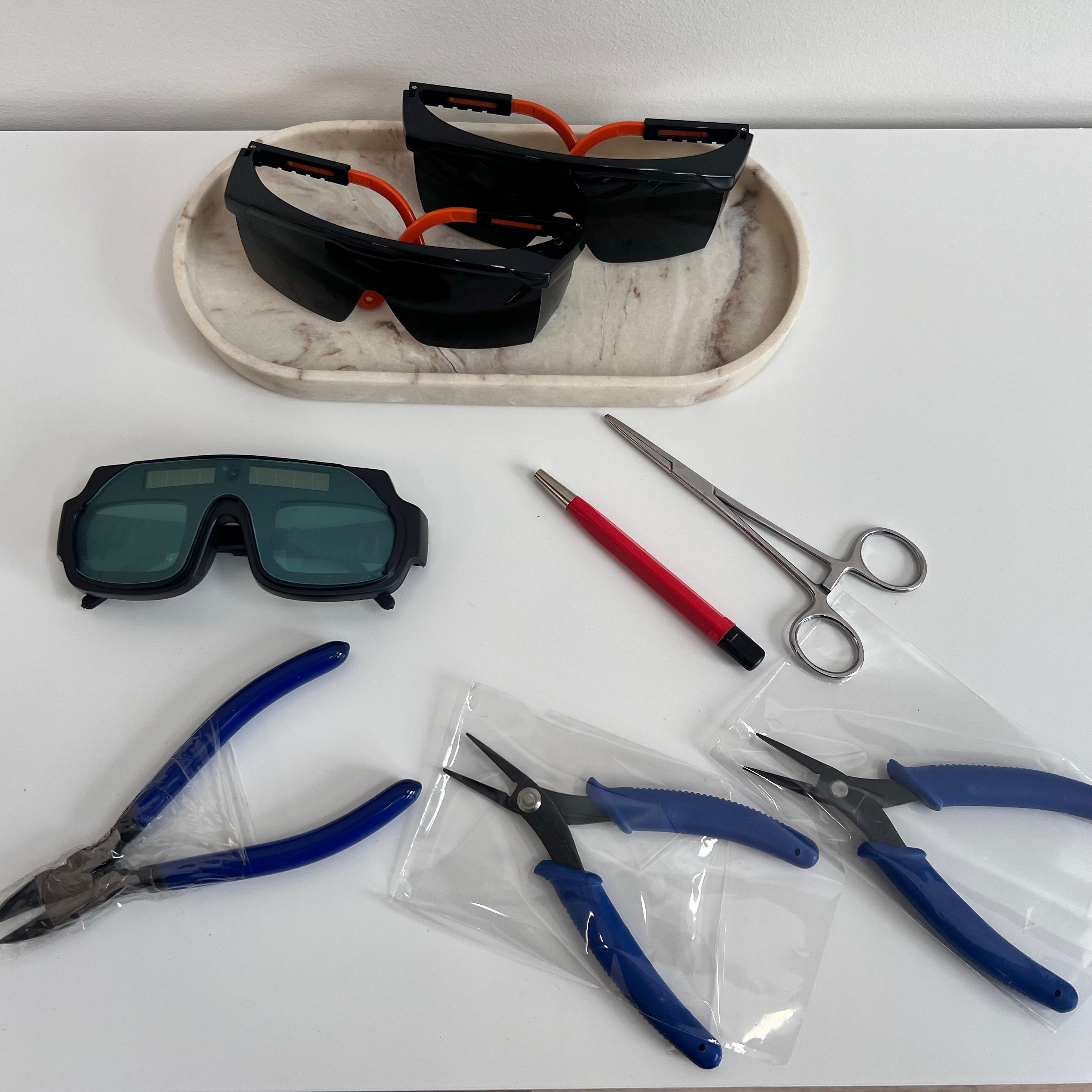 Pair of safety goggles, pliers, scissors, and a pen on a white surface with a marble-patterned tray.